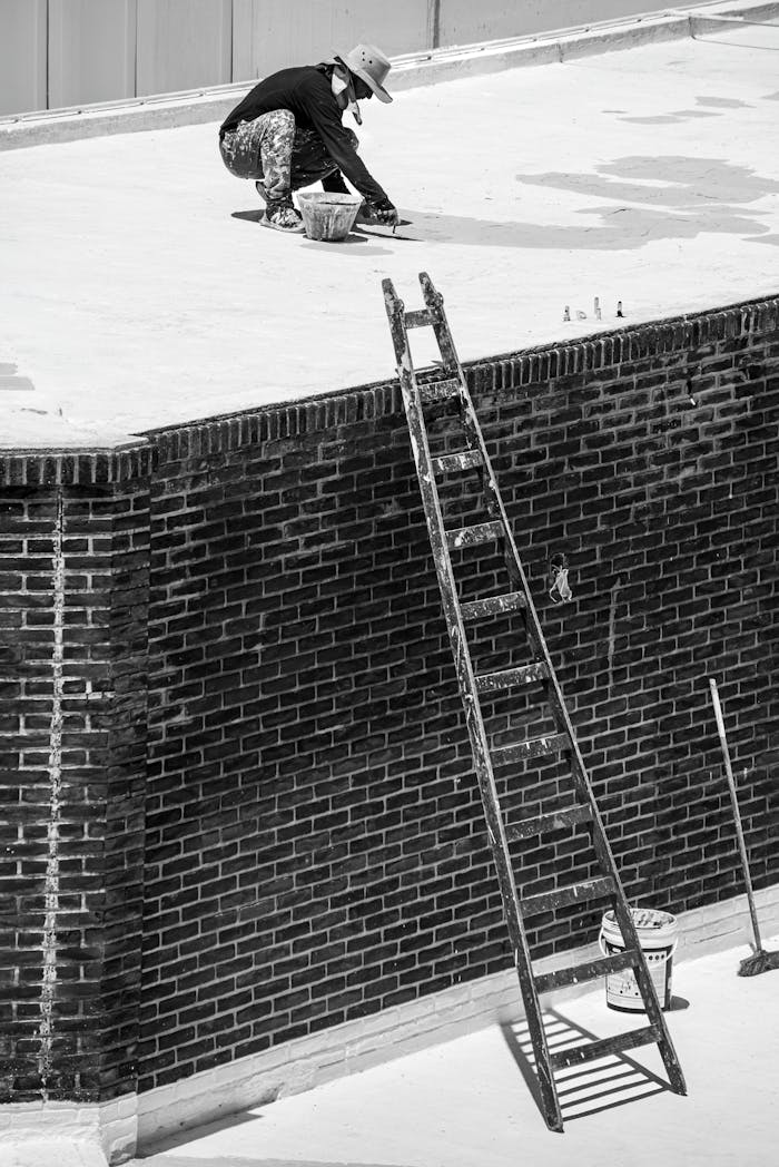 A worker fixing a rooftop while atop a brick building using a ladder.